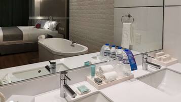 A hotel bathroom with a large mirror, two sinks, a bathtub, and a bed with pillows.