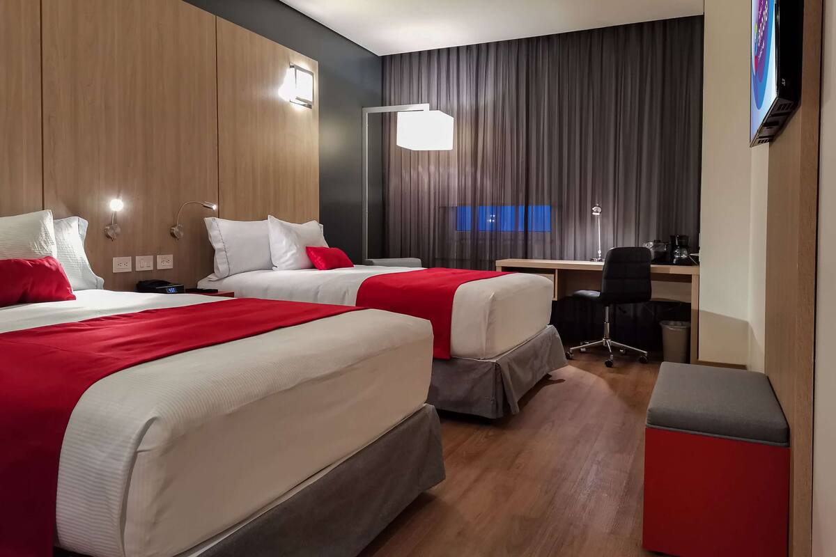 A hotel room with two beds, a desk, a chair, and a TV.