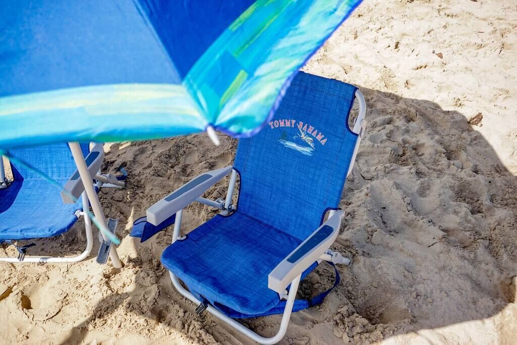 Two beach chairs with a blue umbrella, one with 'TOMMY BARHAM' text, on a sandy beach.