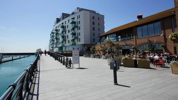 En havnefrontpromenade med en moderne bygning med altaner, en restaurant med udendørs siddepladser og en klar himmel.