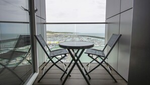 En balkon med et bord og to stole, udsigt over en lysthavn med både og en fjerntliggende kystlinje.