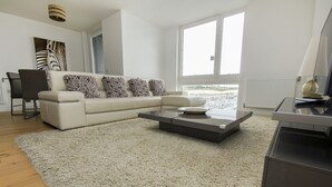 En moderne stue med en beige sofa, et sofabord og et stort vindue med udsigt over byen.