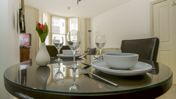 A dining table with a glass top, surrounded by chairs, a white vase with flowers, and empty wine glasses. - No 5 Central by Brighton Holiday Lets (Brighton)