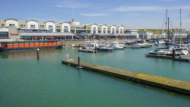 A marina with boats docked, a waterfront building with multiple balconies, and a clear sky.