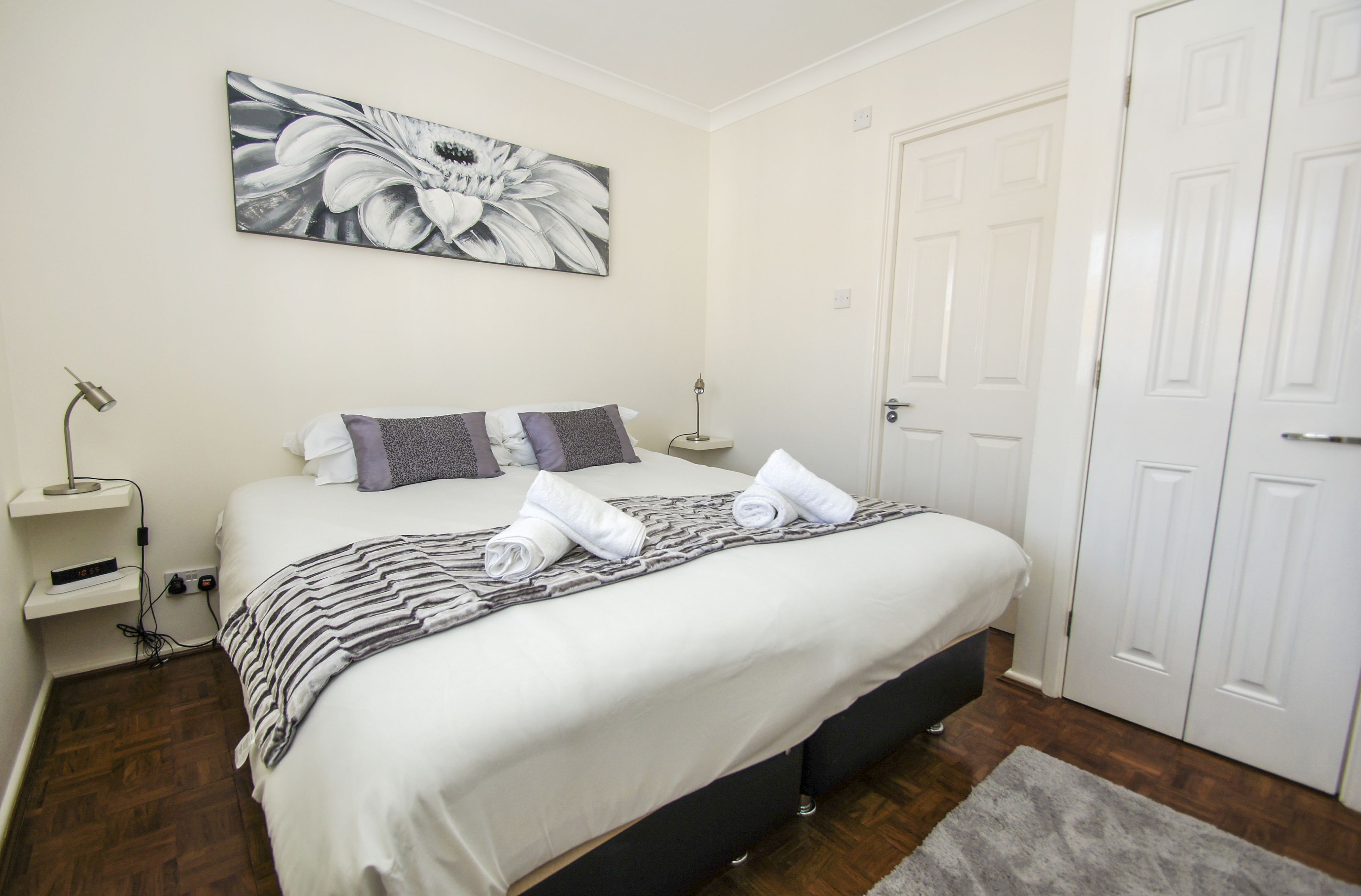 Cama arrumada com travesseiros e cobertor, mesa de cabeceira com abajur e um quadro na parede.
