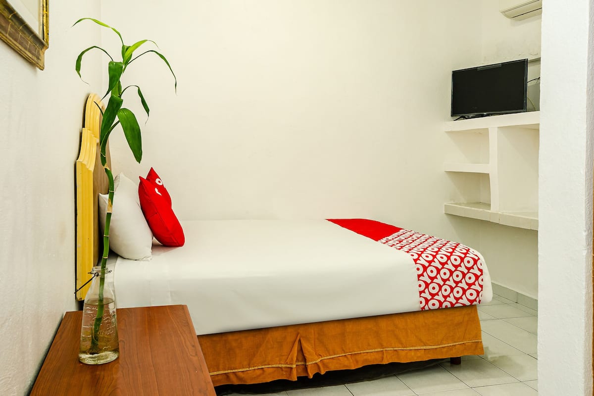 A small, neatly arranged bedroom with a single bed, a red and white patterned blanket, a red pillow, a wooden nightstand with a glass bottle, and a potted plant.