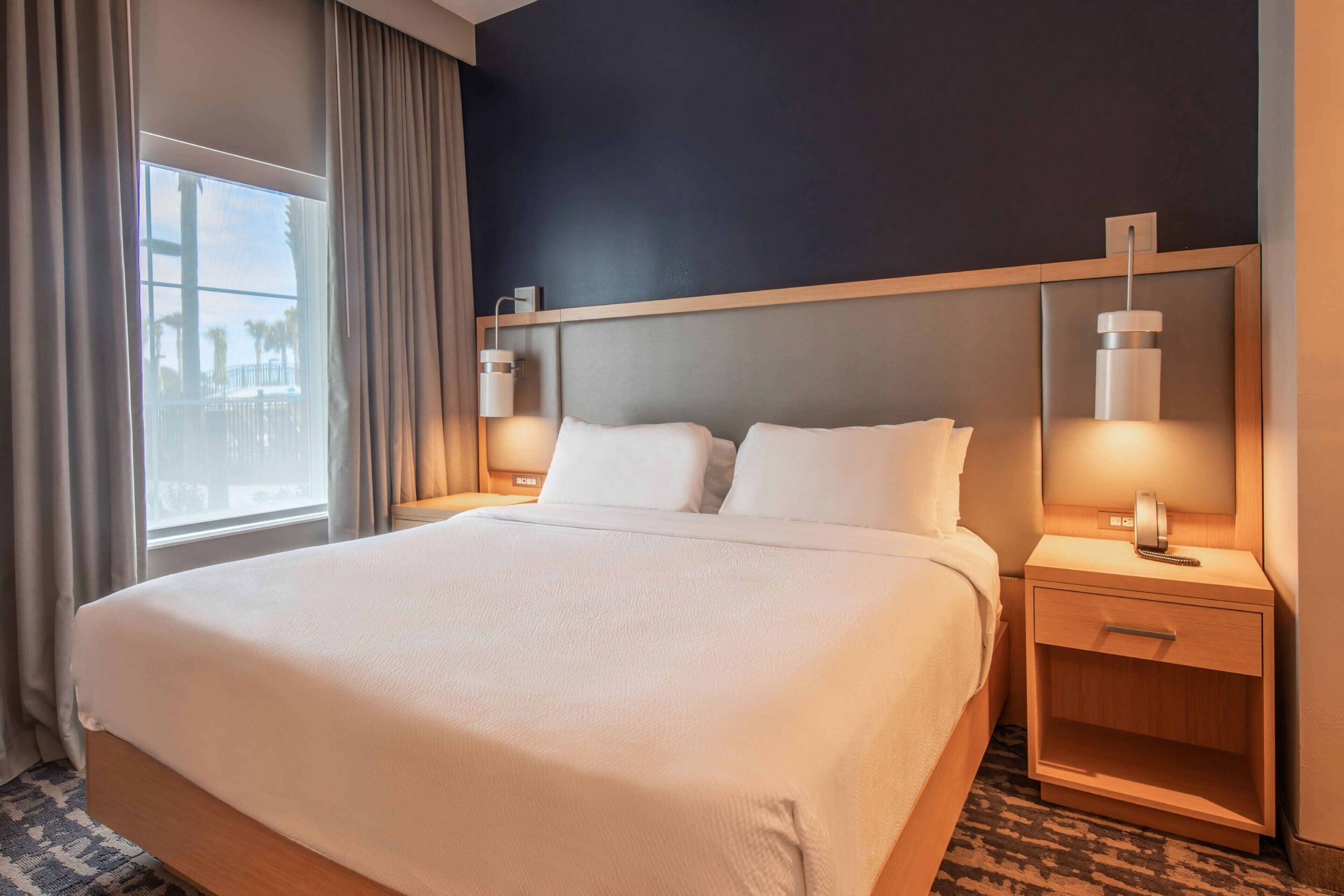 Hotelkamer met een groot bed, een nachtkastje met lamp en uitzicht op palmbomen door het raam.