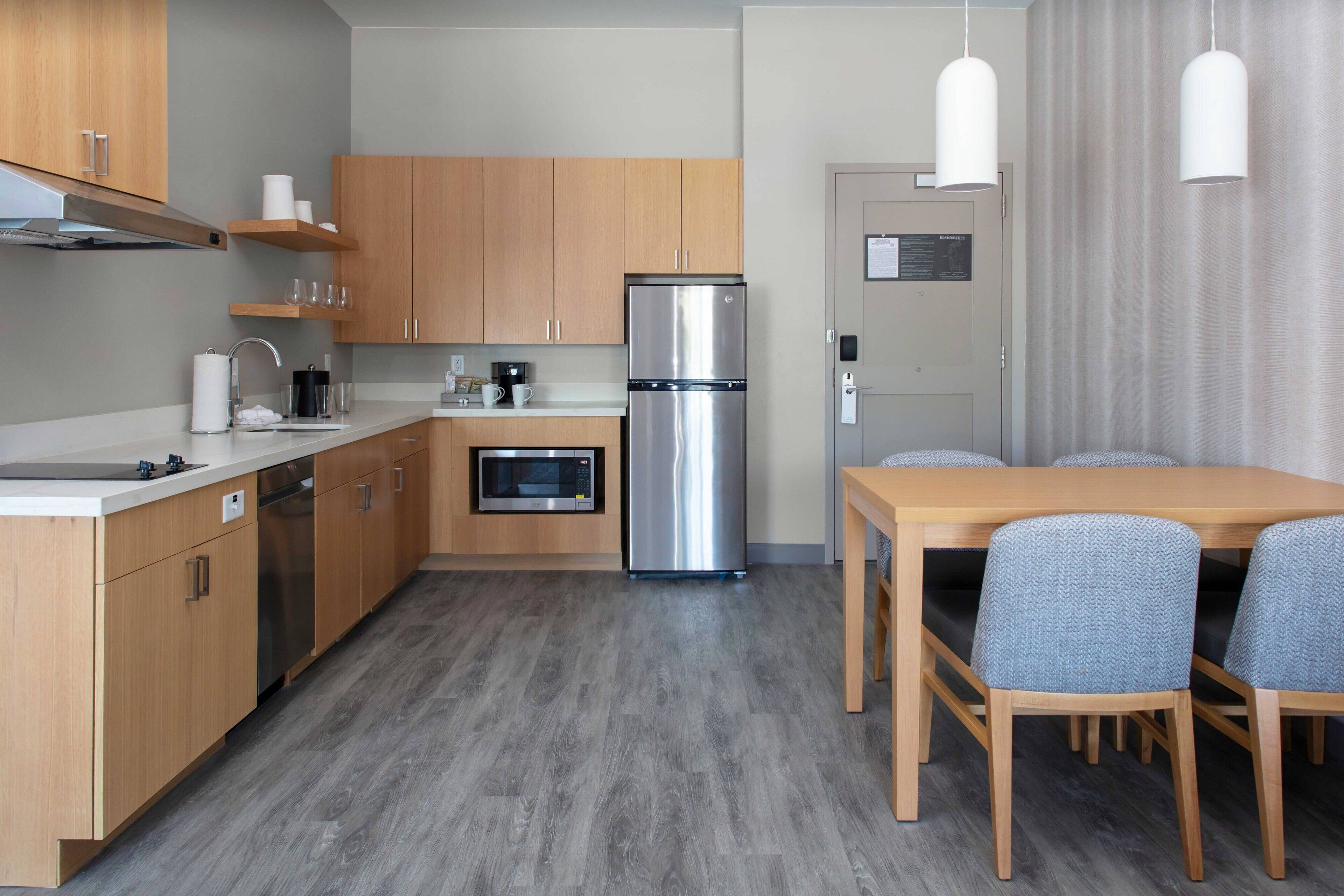 A modern kitchen with wooden cabinets, a stainless steel refrigerator, a microwave, and a dining area with a wooden table and blue chairs.