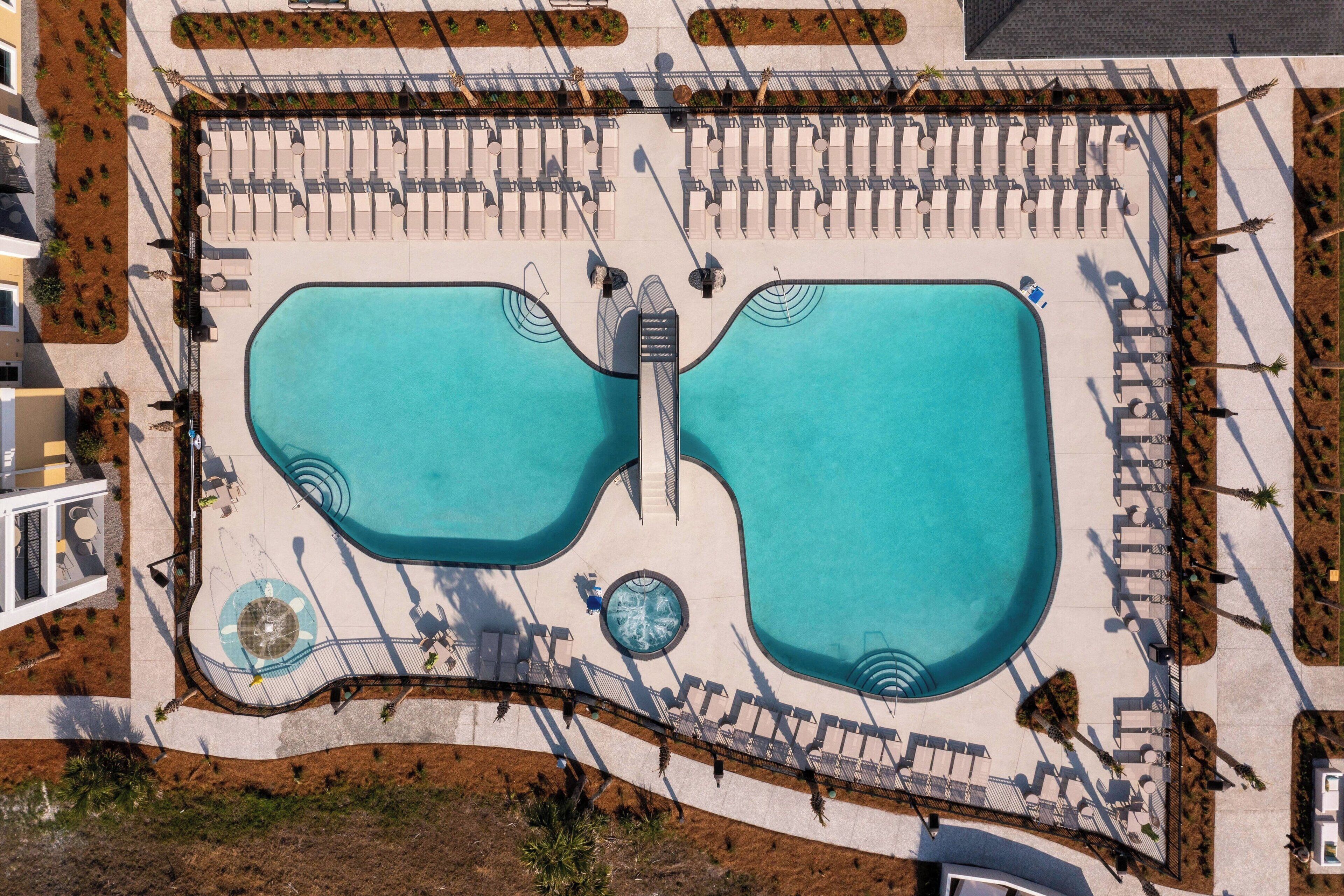Vista aérea de un área de piscina de hotel con dos piscinas grandes, camas de descanso y una fuente central.