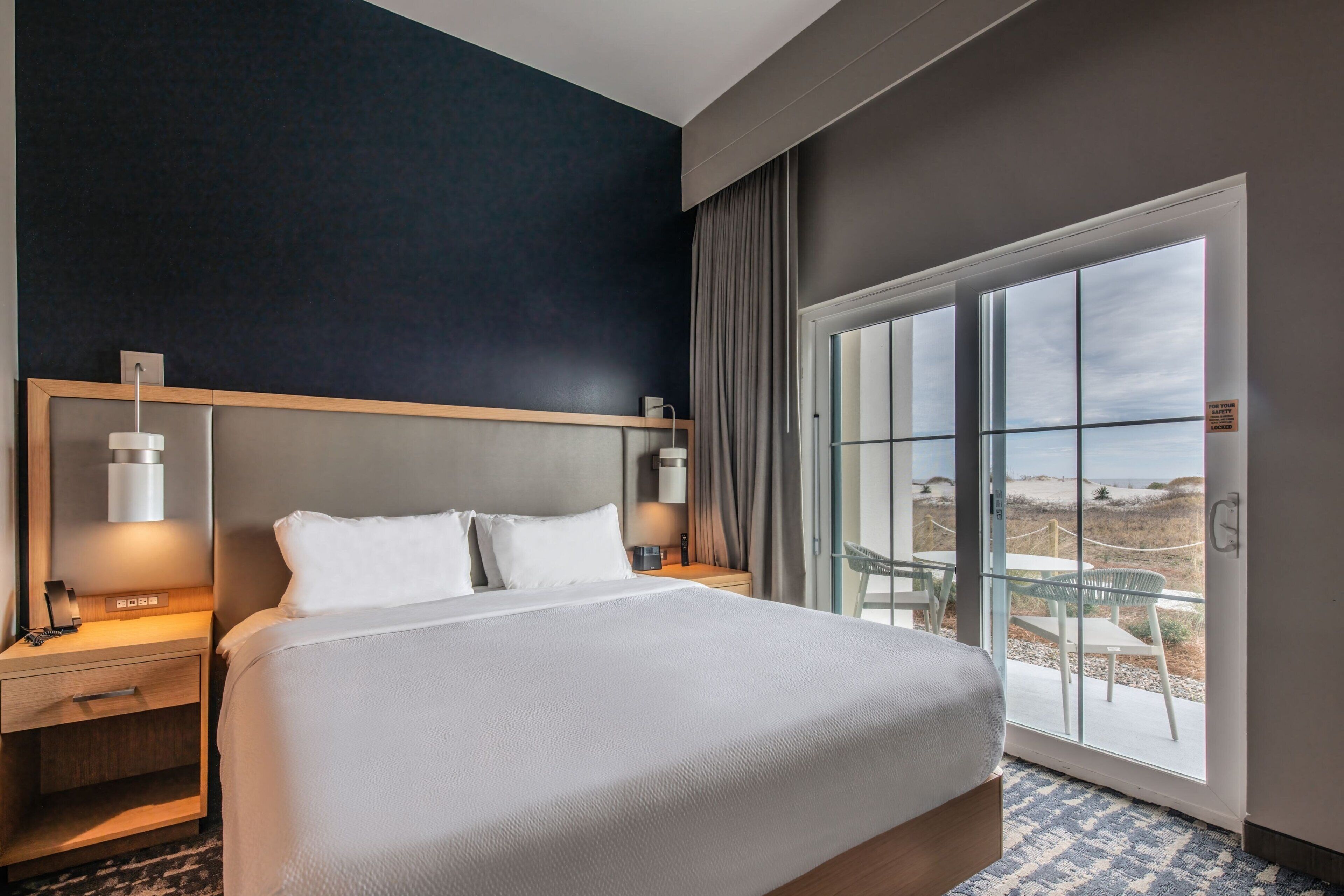 A hotel room with a large bed, bedside lamps, a nightstand, a glass door leading to an outdoor area with a view of the beach.