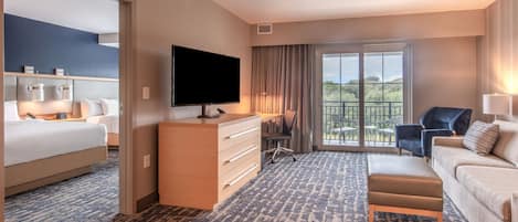 A hotel room with a bed, a desk with a TV, a sofa, and a balcony with a view.