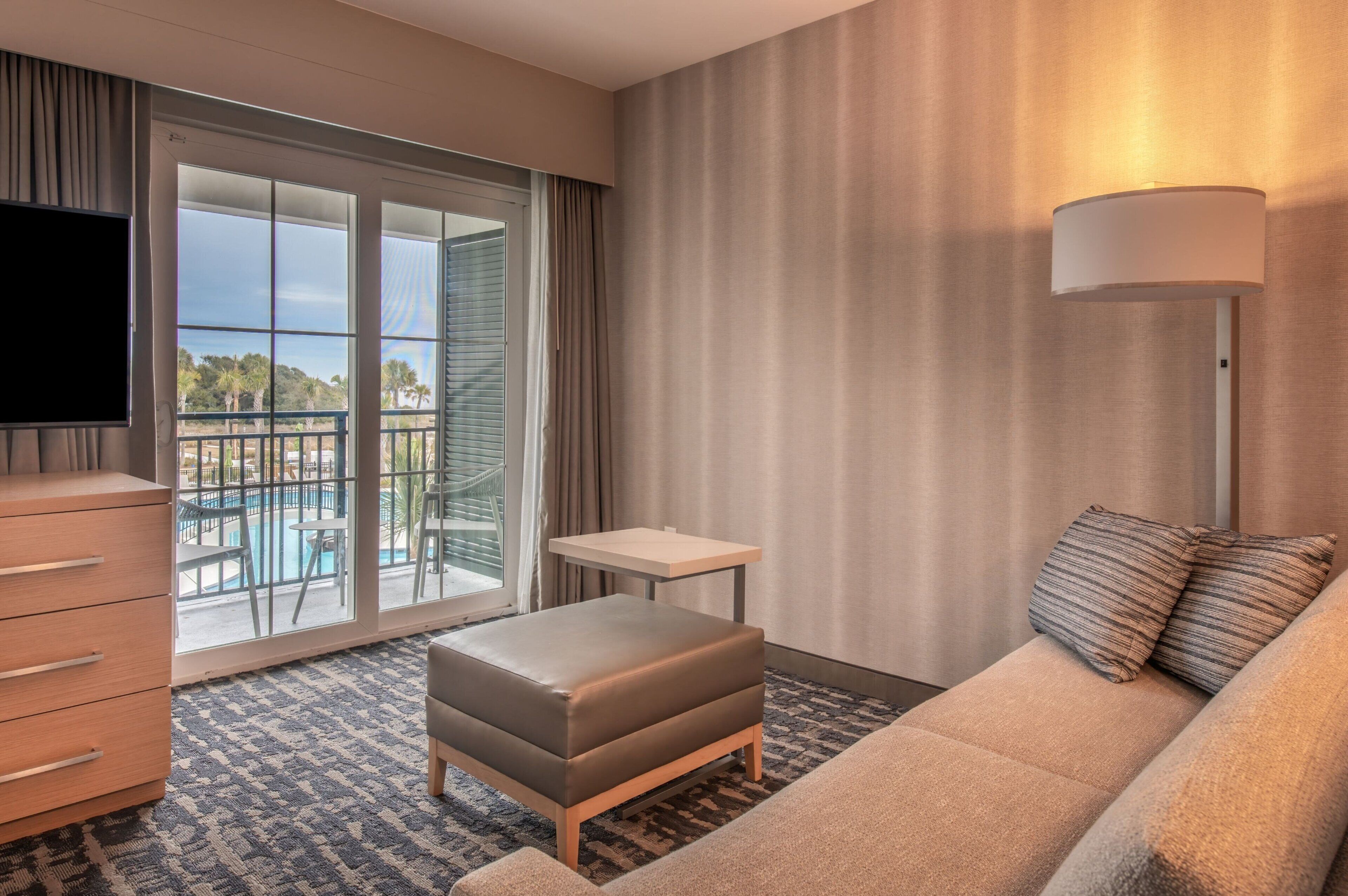 Une chambre d’hôtel avec un canapé, un pouf, une télévision et un balcon donnant sur la piscine.
