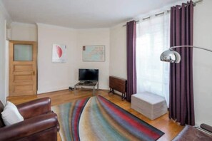 Uma sala de estar com um sofá, televisão, mesa de centro, tapete, janela com cortinas e uma porta.