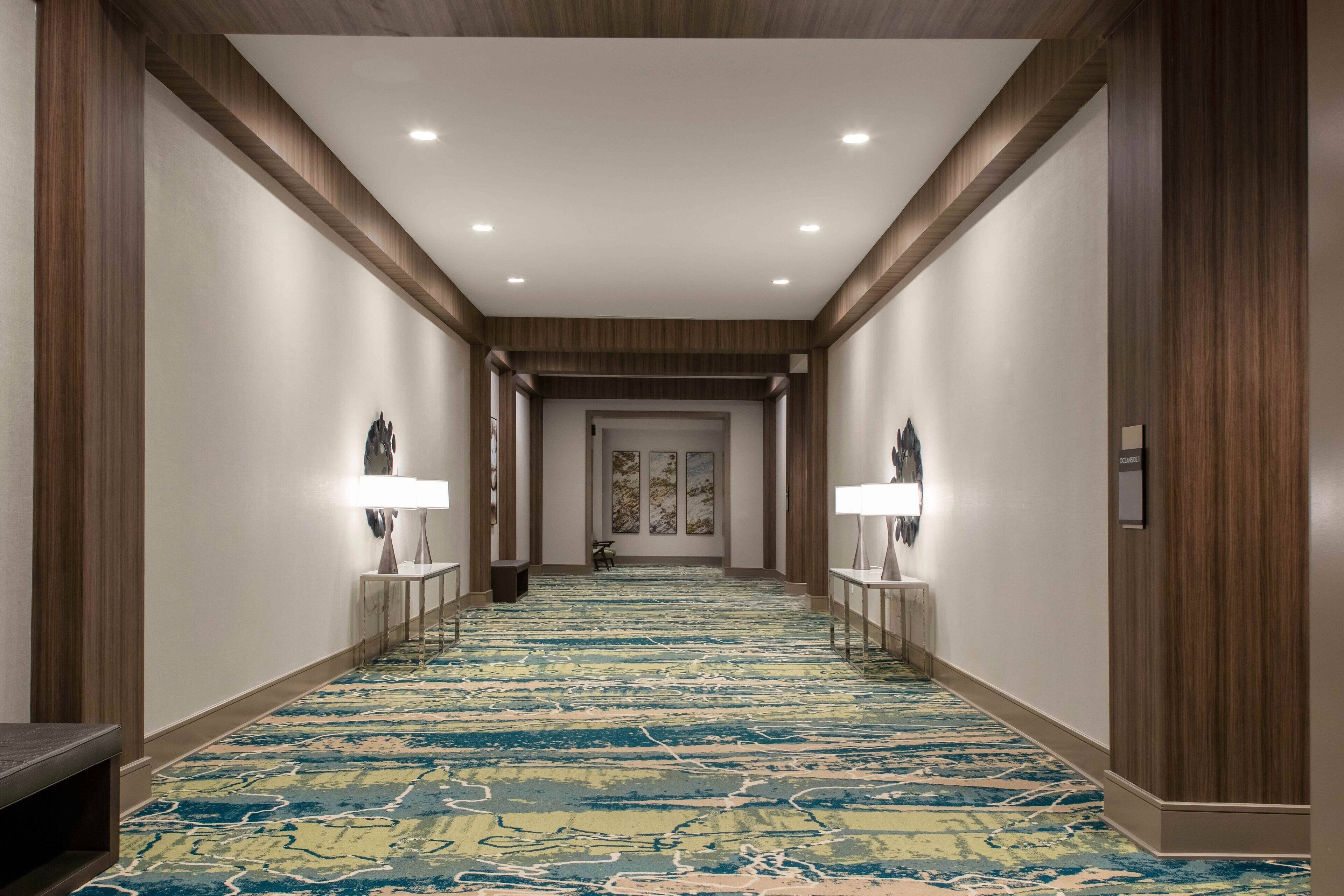 A hotel hallway with a patterned carpet, wooden paneling, and two wall-mounted lamps.