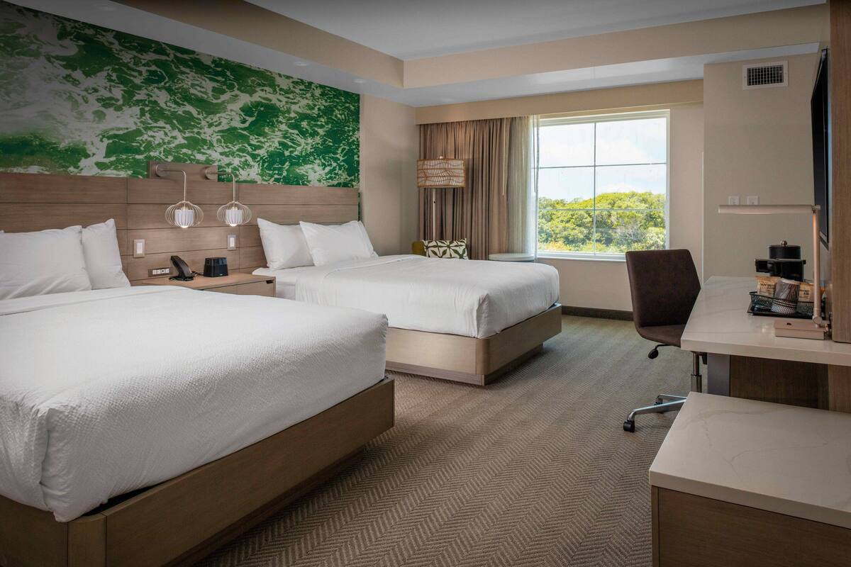 A hotel room with two beds, a desk, a chair, a lamp, and a view of greenery through the window.
