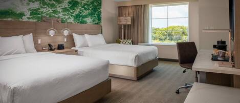 A hotel room with two beds, a desk, a chair, a lamp, and a view of greenery through the window.