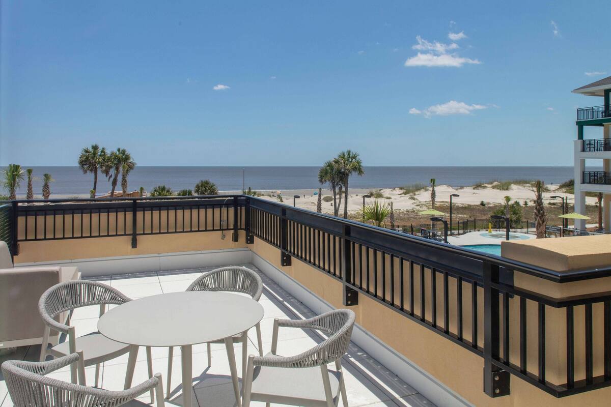 A balcony with a white table and wicker chairs, overlooking a beach with palm trees and a clear sky.