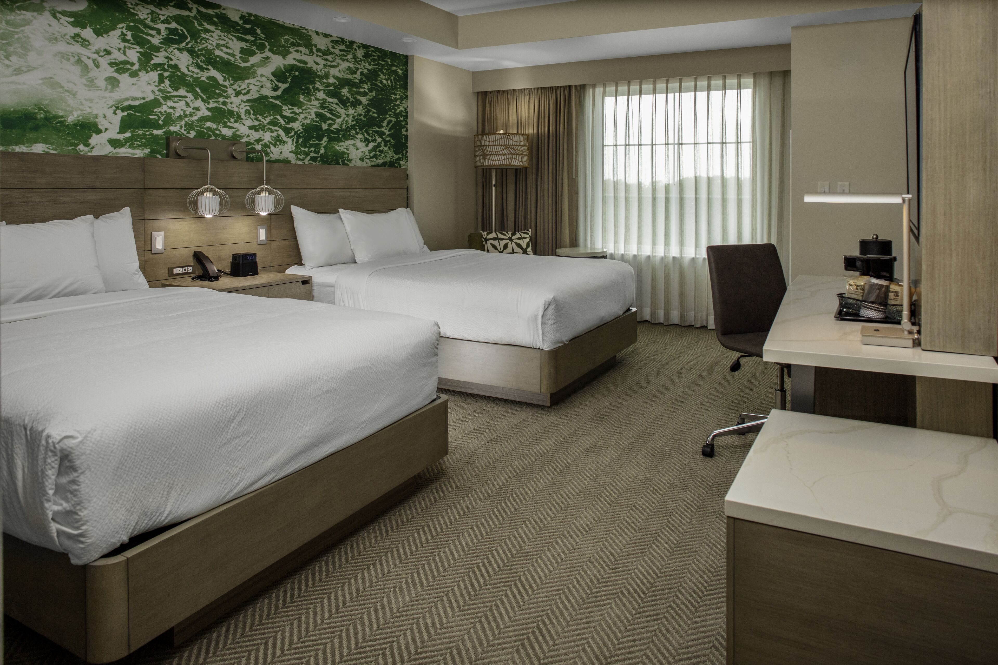 A hotel room with two beds, a desk, a chair, a lamp, and a large window with curtains.