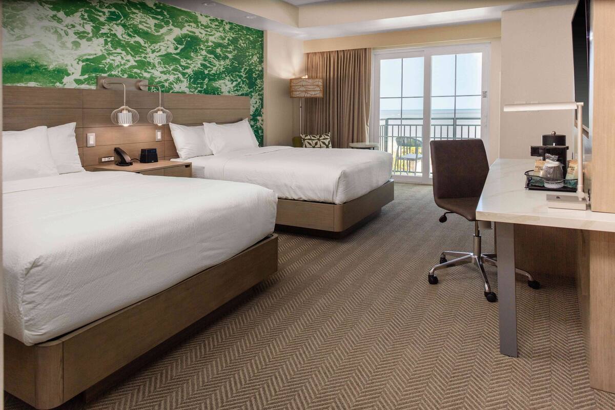 A hotel room with two beds, a desk, a chair, a lamp, and a view of the ocean through the balcony doors.