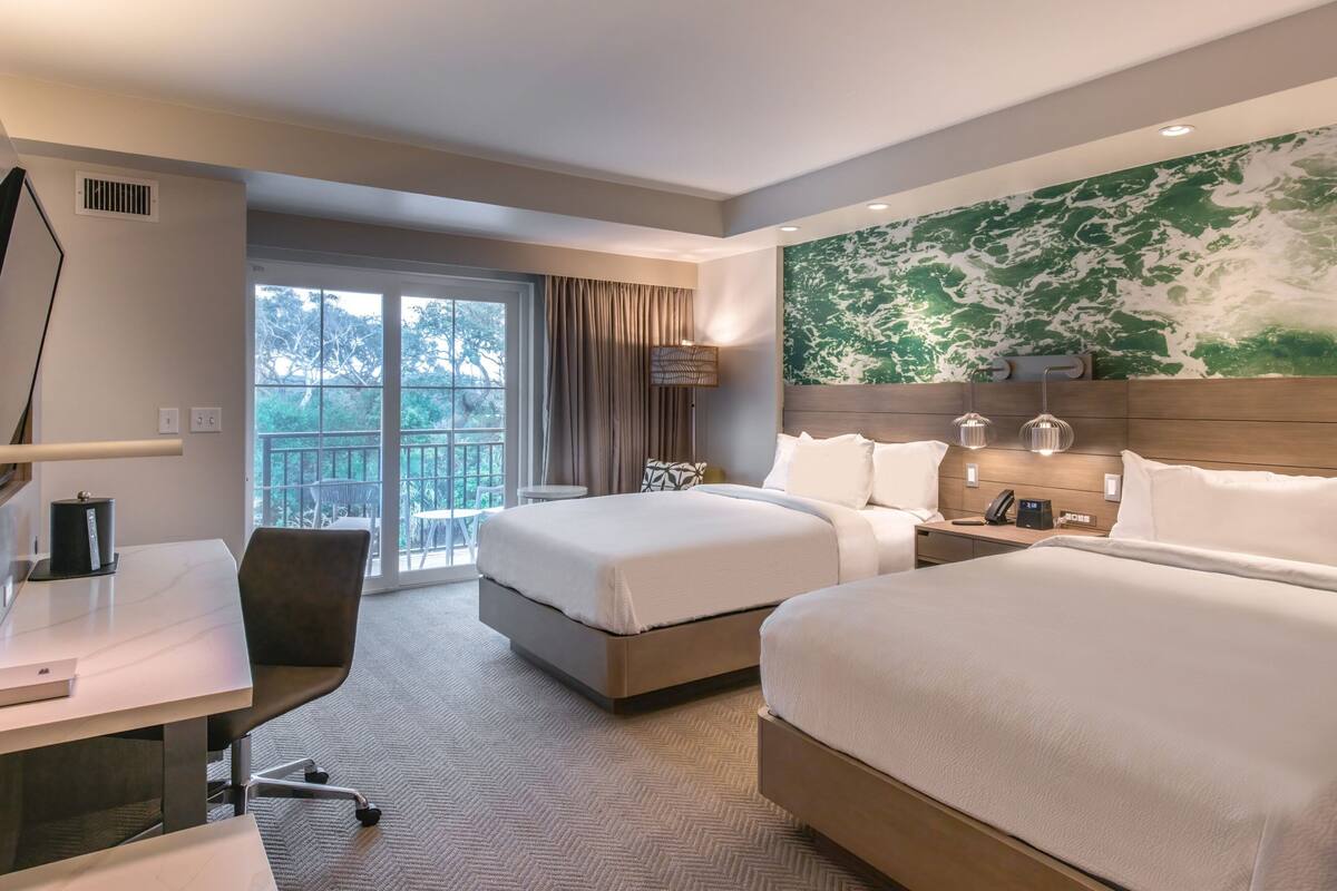A hotel room with two beds, a desk, a chair, a lamp, and a large window with a view of trees.