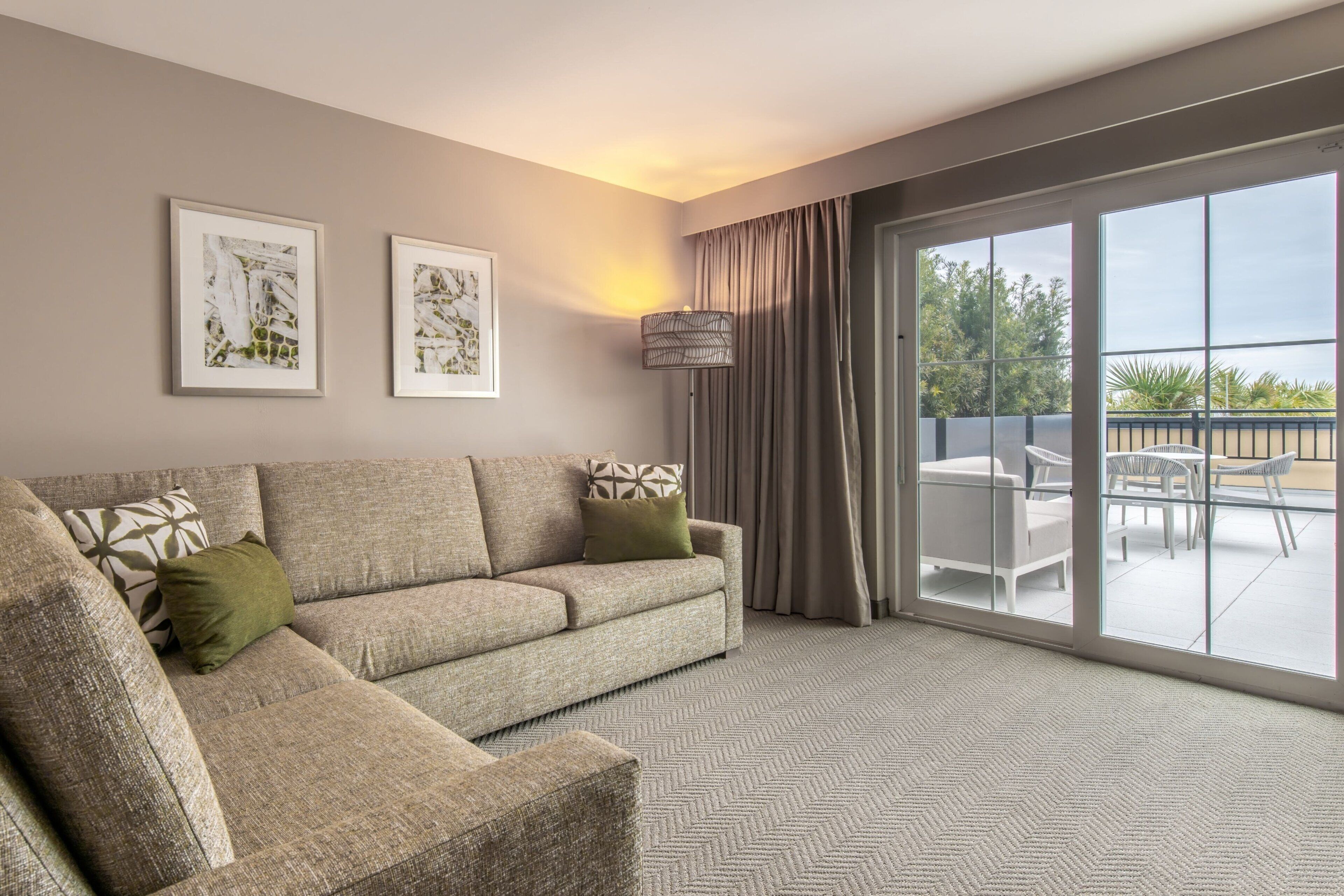 A modern living room with a sofa, two framed artworks, a floor lamp, and a view of the outdoors through large sliding doors.