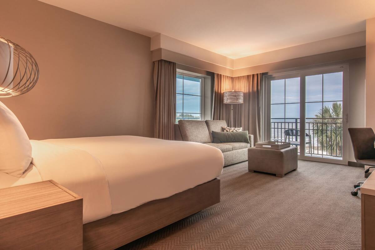 A hotel room with a large bed, a sofa, a desk, and a balcony with a view.
