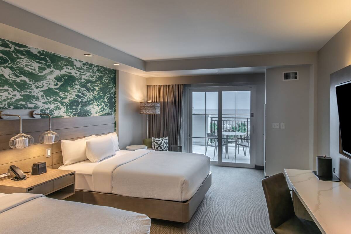 A hotel room with a large bed, a desk, a chair, a TV, a lamp, and a balcony with a view.
