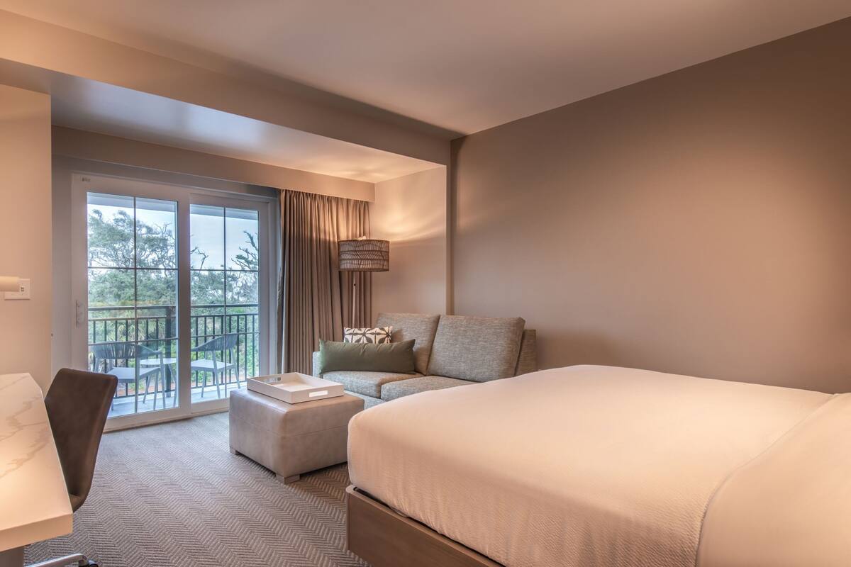 A hotel room with a large bed, a sofa, a small table, and a chair. There is a balcony with a view of trees and a railing.