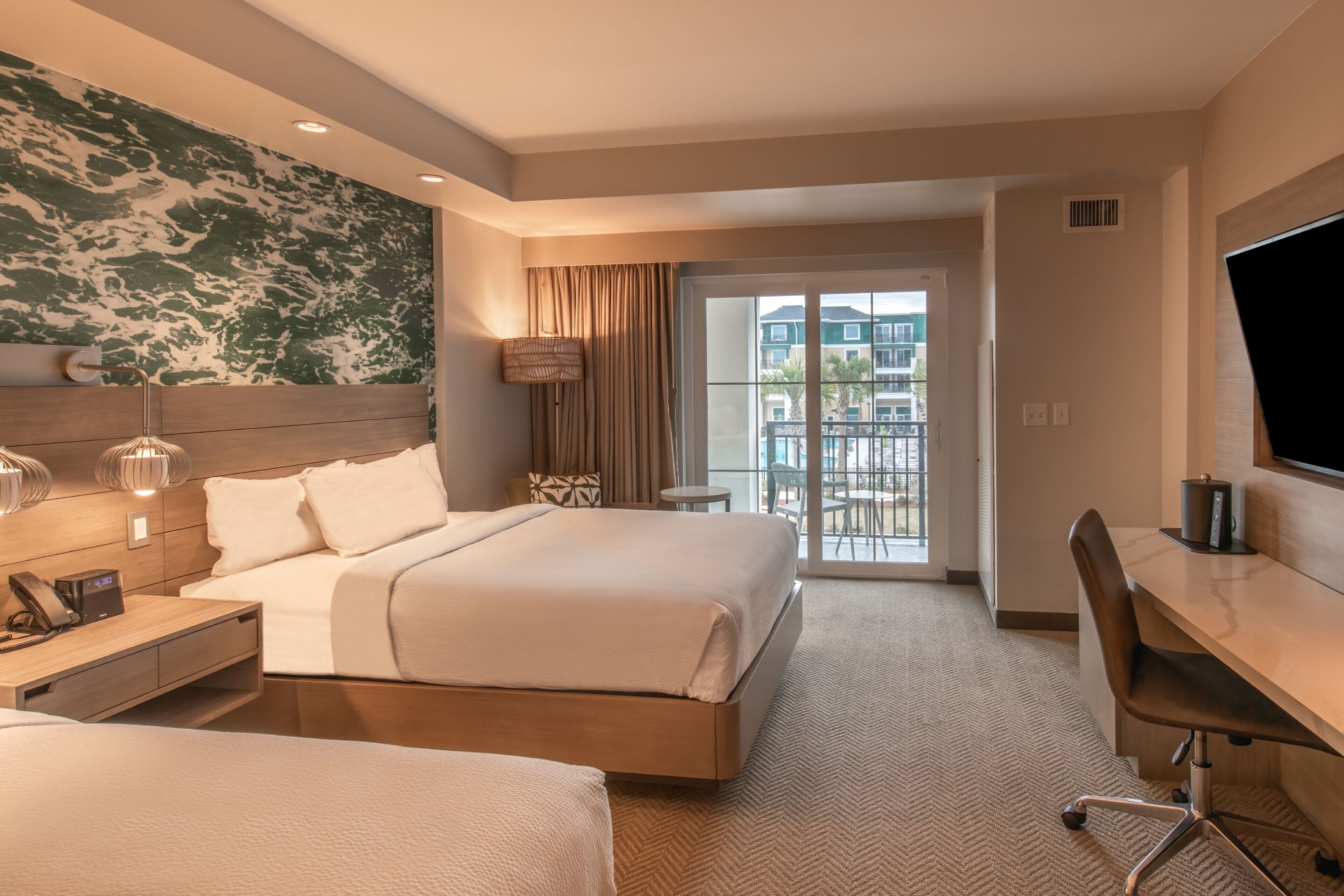 A hotel room with a large bed, a desk, and a TV. There is a balcony with a view of buildings.