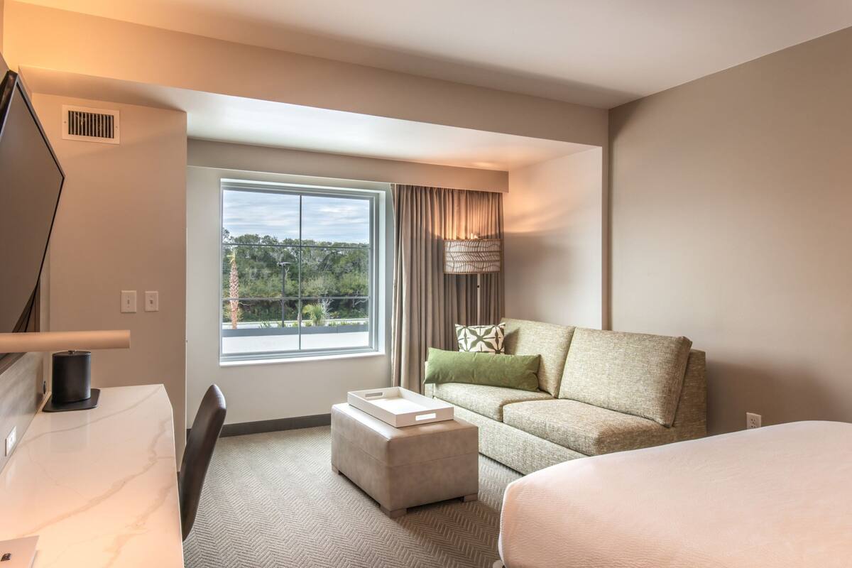 A hotel room with a bed, a sofa, a desk, and a window with a view of greenery.