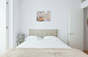 A bedroom with a bed, a nightstand, a lamp, and a wall-mounted picture of a street scene.