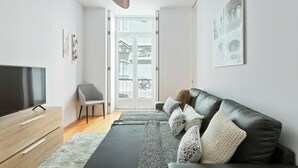 Ein modernes Wohnzimmer mit einem Flachbildfernseher, einem grauen Sessel, einem schwarzen Ledersofa mit gemusterten Kissen und einem großen Fenster mit Blick auf einen Balkon.