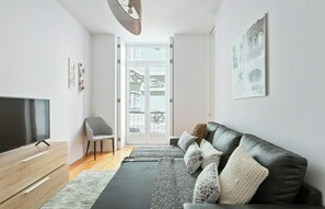 Ein modernes Wohnzimmer mit einem Flachbildfernseher, einem grauen Sessel, einem schwarzen Ledersofa mit gemusterten Kissen und einem großen Fenster mit Blick auf einen Balkon.