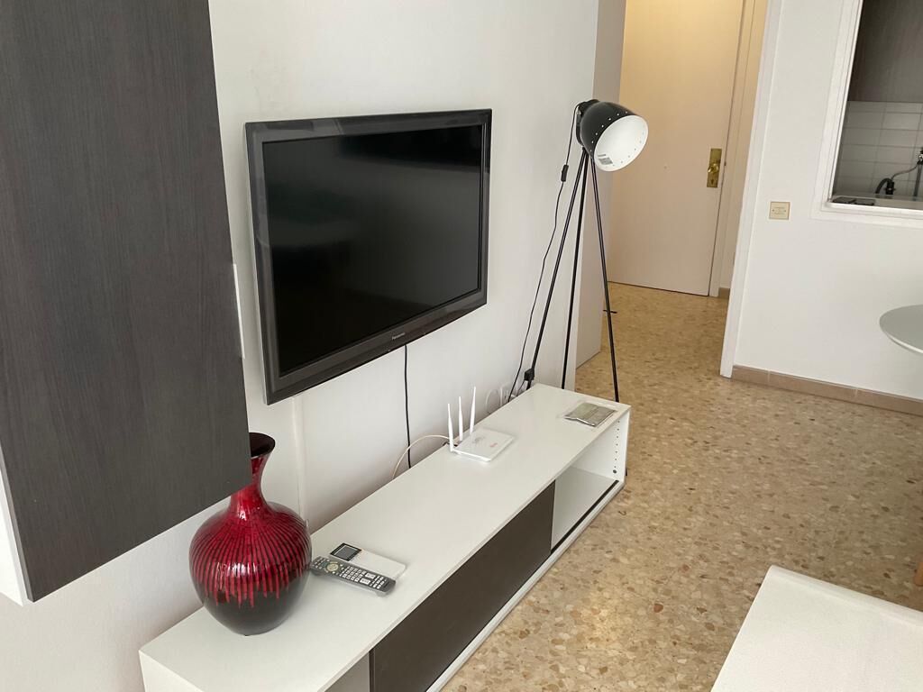 A modern living room with a flat-screen TV mounted on the wall, a white console table with a red vase and a remote control, and a floor lamp.