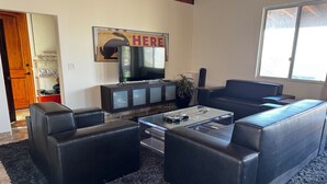 A living room with black leather sofas, a glass coffee table, a TV on a cabinet, and a 'HERE' sign on the wall. - Amazing Retreat with Unbelievable Views in Malibu (Malibu)