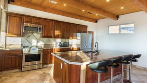 A modern kitchen with wooden cabinets, stainless steel appliances, a large island with bar stools, and a view of the outside through a window. - Amazing Retreat with Unbelievable Views in Malibu (Malibu)