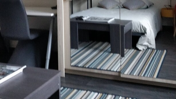 Ein Hotelzimmer mit Bett, Schreibtisch, Stuhl, einem an der Wand montierten Fernseher und einem kleinen Regal mit Gegenständen.