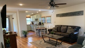 A spacious living area with a dark leather sofa, a coffee table, and a dining table with chairs. A kitchen with light-colored cabinets and a ceiling fan is visible in the background.