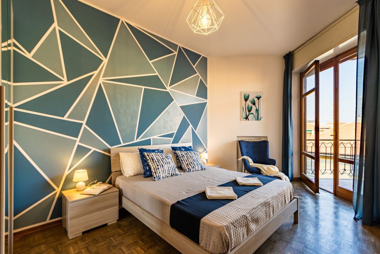Une chambre moderne avec un motif géométrique sur le mur, un lit recouvert d’une courtepointe bleue et blanche, une table de chevet avec une lampe, et une vue sur l’extérieur grâce à une grande fenêtre.