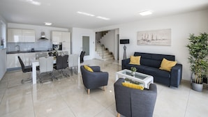 Ein modernes Wohnzimmer mit einem dunklen Sofa, zwei Sesseln, einem Couchtisch und einem Essbereich mit weißem Tisch und Stühlen.
