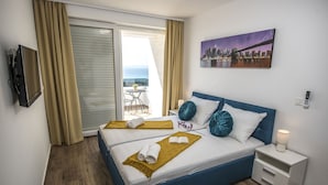 Ein Hotelzimmer mit Bett, Nachttisch, Fernseher, einem Gemälde und Blick auf Stadt und Meer.