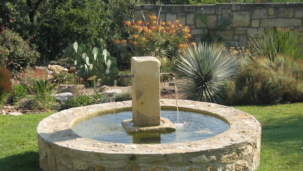Een ronde stenen fontein met een centrale pilaar, omgeven door een gazon en verschillende woestijnplanten.