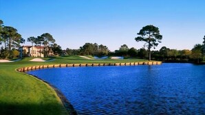 Ein Golfplatz mit einem Wasserhindernis, Sandbunkern und einem Clubhaus im Hintergrund.