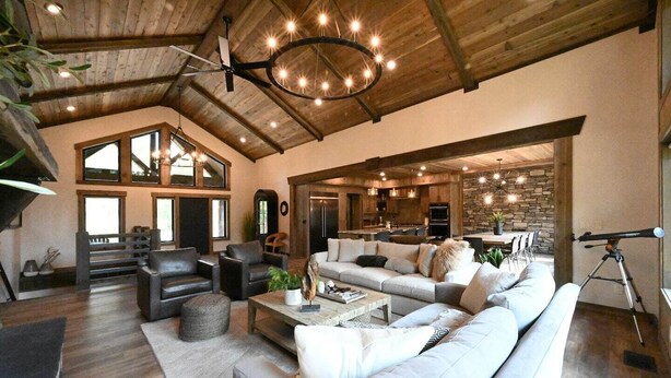 Un'ampia zona giorno con soffitto alto, travi in legno e un grande lampadario. C'è un'area salotto con divani e tavolino, e una cucina sullo sfondo.