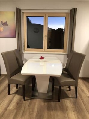 Un coin repas avec une table blanche et quatre chaises, une fenêtre avec des rideaux et un petit objet décoratif posé sur la table.