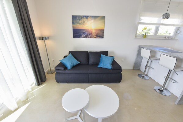 A modern living area with a sofa, two round tables, a kitchen area with bar stools, and a large window with curtains. - Mare Deluxe Apartment with Pool and Terrace (Novalja)