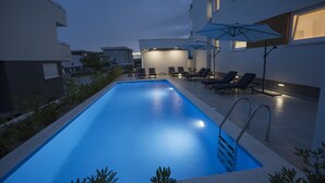 Piscina com espreguiçadeiras e guarda-sóis, rodeada por edifícios.
