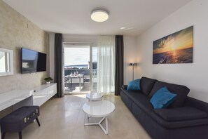 A modern living room with a flat-screen TV, a black sofa with blue cushions, a white coffee table, and a painting of a sunset. - Mare Deluxe Apartment with Pool and Terrace (Novalja)