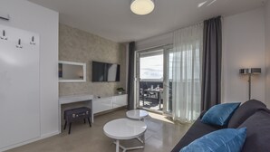 Uma sala de estar moderna com um sofá, duas mesas brancas, uma televisão de tela plana e uma porta de vidro deslizante que dá para uma varanda.