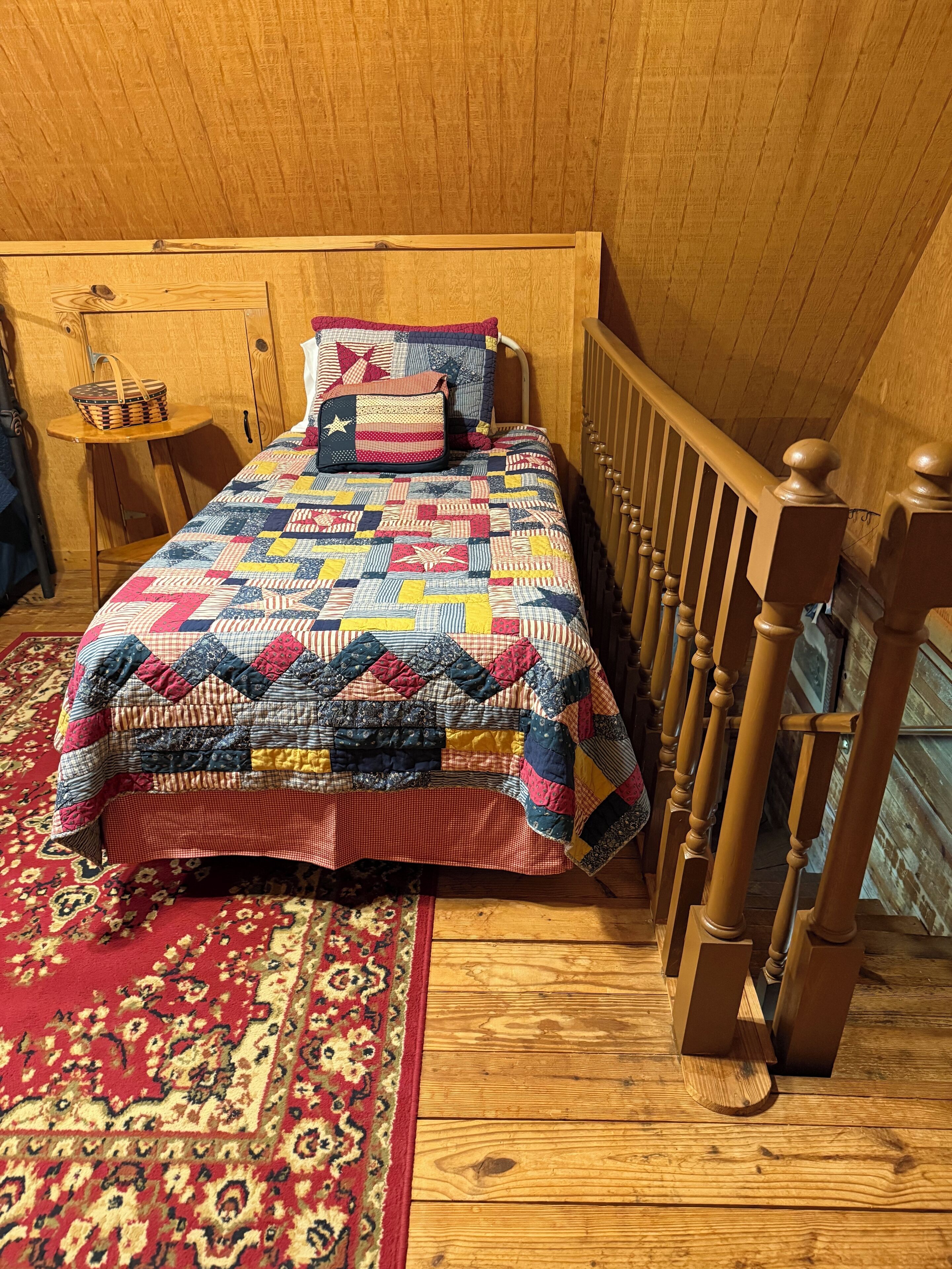 Une chambre avec un lit en bois, une couette rouge et bleue, une tête de lit en bois, une barrière en bois et un tapis à motifs.